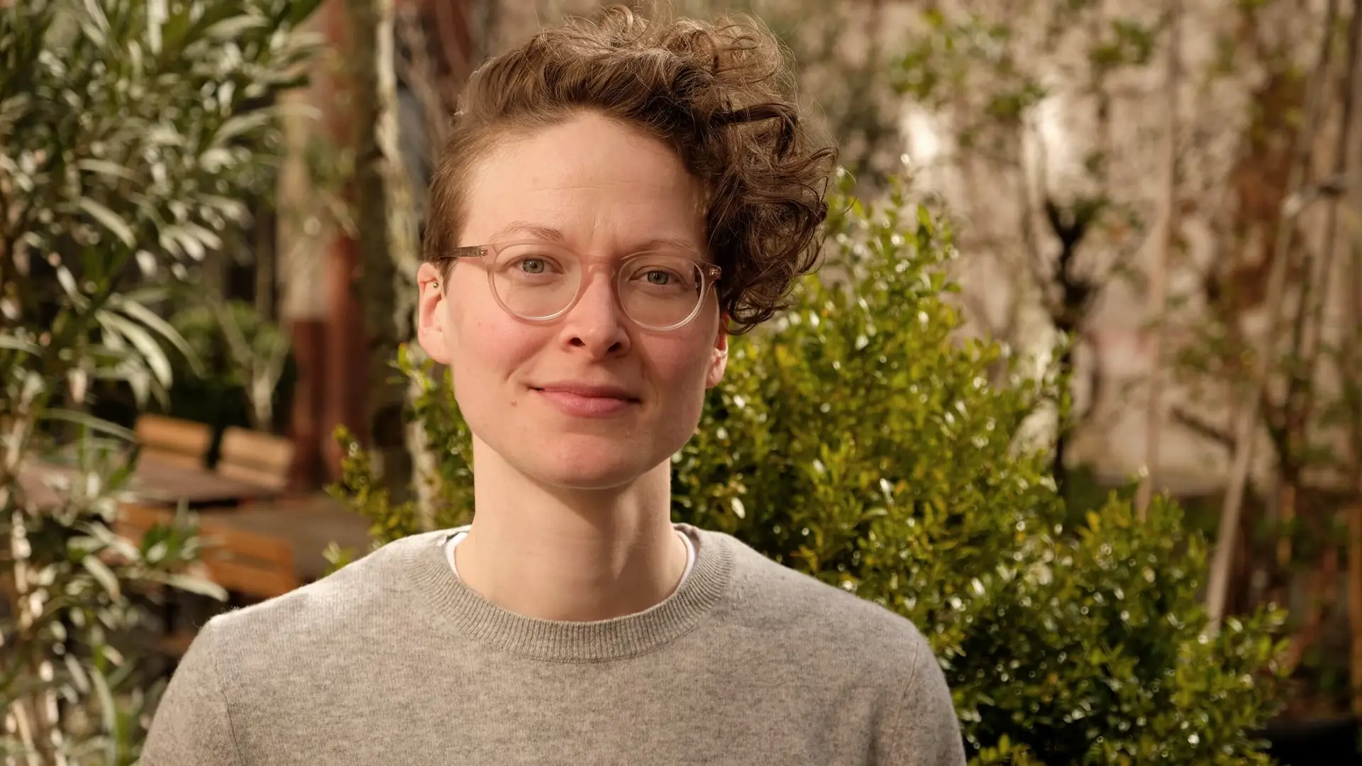 Portrait Lena Wigger, eine Frau mit kurzen blonden Locken, hellblauen Augen und einer Brille.