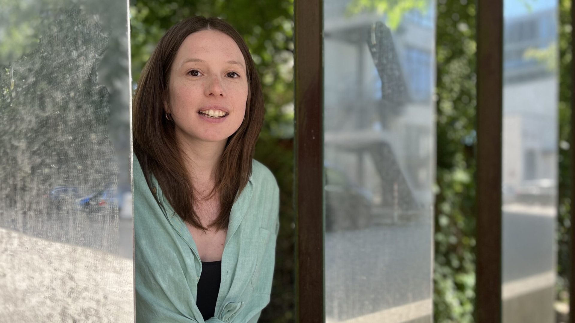 Portrait Foto von Louise Fischer. Louise hat braune lange Haare, einen Mittelscheitel, braune Augen und helle Haut. Sie trägt eine Aquamarinblaue Bluse, die am Bauch geknotet ist und dazu ein schwarzes Top. Sie steht zwischen Spiegelsäulen.