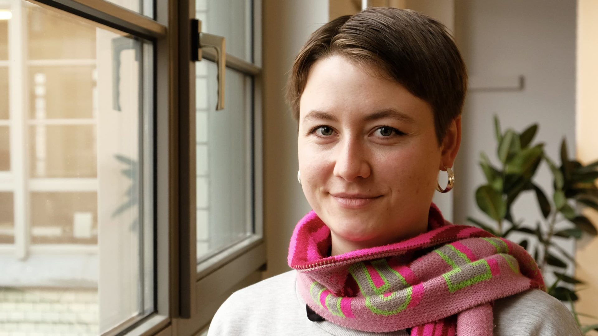 Portrait Leonie Wetter. Eine Frau mit kurzen bruanen Haaren, pinkgrünem Schal und grauem Pullover steht an einem Fenster,